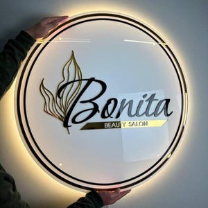 Może przedstawiać: Okrągły, podświetlany szyld salonu piękności. Szyld zawiera słowo "Bonita" w stylowej czarnej czcionce, z napisem "BEAUTY SALON" poniżej. Złoty wz&oacute;r liści i złote akcenty obramowują białe tło.