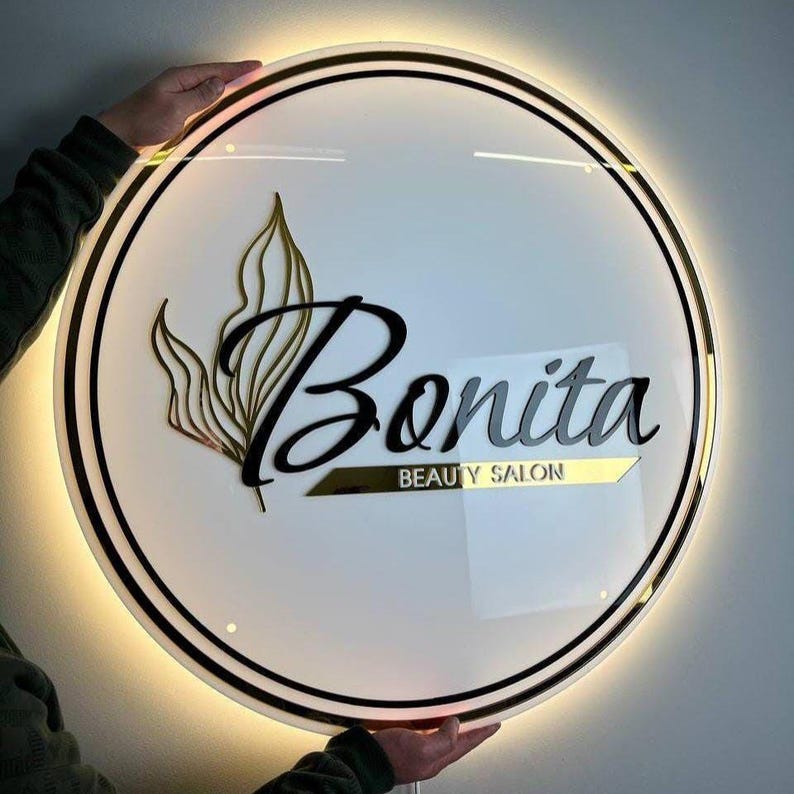Może przedstawiać: Okrągły, podświetlany szyld salonu piękności. Szyld zawiera słowo "Bonita" w stylowej czarnej czcionce, z napisem "BEAUTY SALON" poniżej. Złoty wz&oacute;r liści i złote akcenty obramowują białe tło.