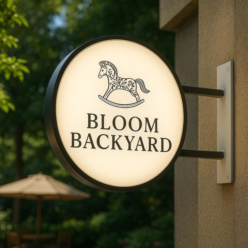 K&ouml;nnte beinhalten: Ein rundes, beleuchtetes Schild mit dem Text "BLOOM BACKYARD" und einer Schaukelpferd-Illustration. Das Schild hat einen schwarzen Rahmen und ist an einer beigefarbenen Wand mit einer wei&szlig;en Halterung befestigt. Der Hintergrund zeigt Gr&uuml;n und einen Sonnenschirm.