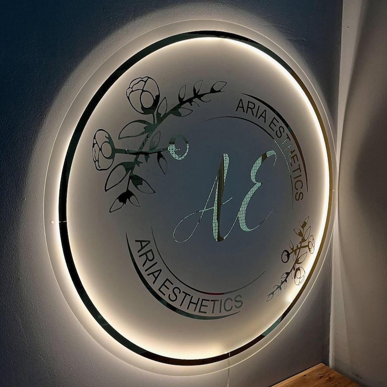 Może przedstawiać: Okrągły znak z napisem "ARIA ESTHETICS" w zakrzywionej czcionce na krawędzi. Środek zawiera litery "AE" w ozdobnym skrypcie, otoczone kwiatowymi wzorami. Znak jest podświetlony ciepłym, miękkim światłem.