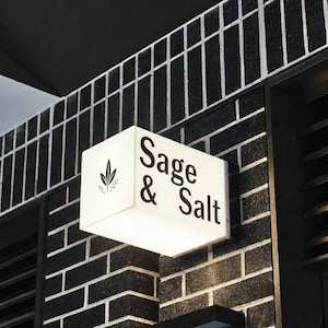May include: A white, illuminated sign with the words "Sage & Salt" in black lettering. A small leaf design is on the left side. The sign is mounted on a black brick wall. The sign is square and has a modern design.