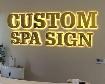 May include: A wall-mounted sign featuring three-dimensional gold-colored letters that spell out CUSTOM SPA SIGN. The letters are illuminated from behind with a warm glow, creating a halo effect against the plain, neutral-colored wall surface.