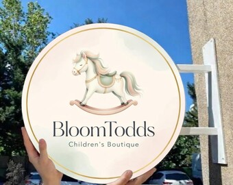 Könnte beinhalten: Rundes Schild für die Kinderboutique BloomTodds mit einer Illustration eines Schaukelpferds in Pastellfarben. Das Schild hat einen goldenen Rand und wird von einer weißen Halterung gehalten. Der Text "BloomTodds" und "Children's Boutique" sind zu sehen.