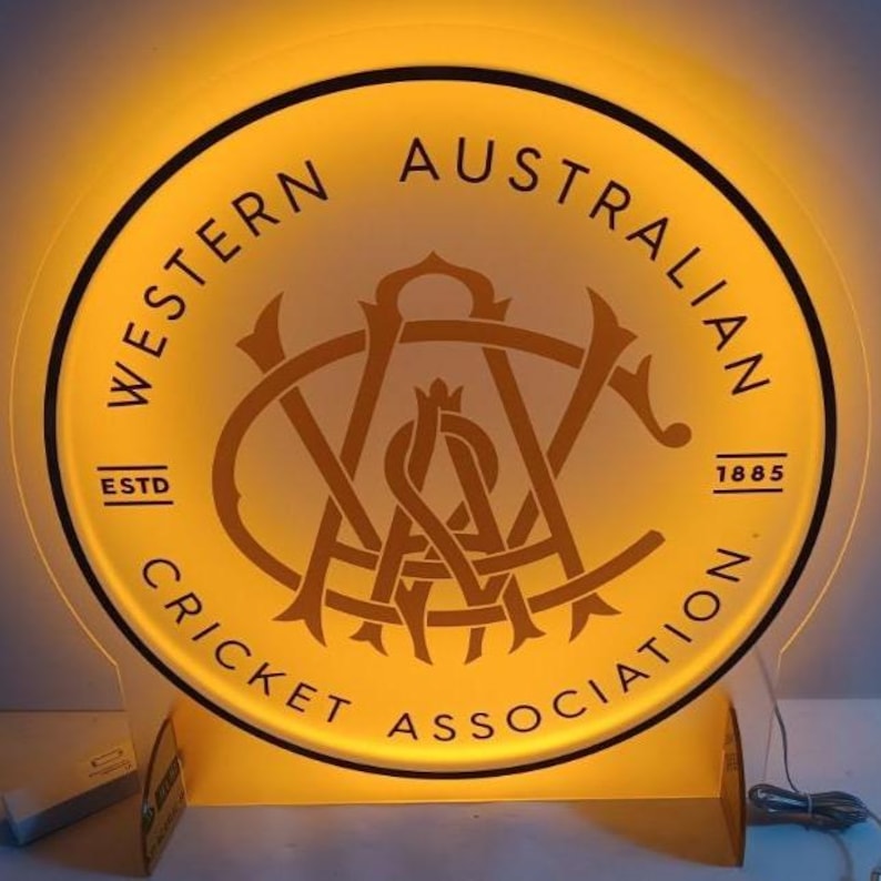 Może przedstawiać: Okrągły, podświetlany znak z napisem "WESTERN AUSTRALIAN CRICKET ASSOCIATION" w okręgu. Znak zawiera centralne logo i rok "1885". Znak emituje ciepłą, ż&oacute;łtą poświatę.