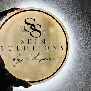 Może przedstawiać: Okrągły, podświetlany szyld z czarnym napisem "SS SKIN SOLUTIONS by 2 divas". Znak ma złoty środek z czarną obw&oacute;dką i białym światłem na krawędzi. Logo zawiera splecione litery "SS" i akcenty liści.
