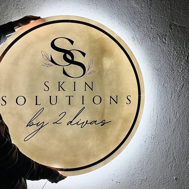 Może przedstawiać: Okrągły, podświetlany szyld z czarnym napisem "SS SKIN SOLUTIONS by 2 divas". Znak ma złoty środek z czarną obw&oacute;dką i białym światłem na krawędzi. Logo zawiera splecione litery "SS" i akcenty liści.