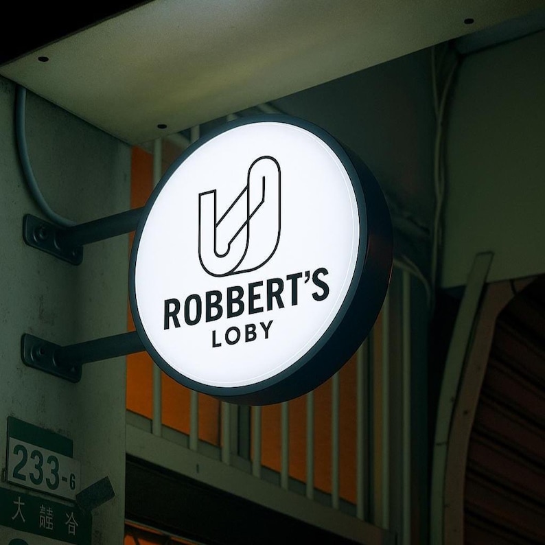 K&ouml;nnte beinhalten: Ein rundes, beleuchtetes Schild mit dem Text "ROBBERT'S LOBY" in Schwarz, unter einer stilisierten schwarzen Grafik. Das Schild ist an einer dunklen Metallhalterung an einer hellen Wand befestigt. Ein gr&uuml;nes Schild mit Zahlen ist sichtbar.