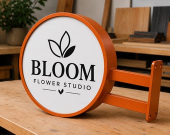Könnte beinhalten: Ein rundes, orangefarbenes Wand-Metallschild mit weißer Vorderseite und schwarzer Aufschrift BLOOM FLOWER STUDIO, ergänzt durch ein schwarzes Blattsymbol über dem Text und ein kleines Blatt zentriert unter den Worten FLOWER STUDIO.