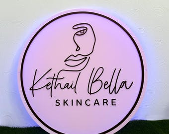 Könnte beinhalten: Ein runder, rosafarbener Aufsteller mit schwarzem Rand und dem Text "Kethail Bella SKINCARE". Das Schild zeigt eine minimalistische Strichzeichnung eines Gesichts und eignet sich für ein Schönheits- oder Hautpflegeunternehmen.