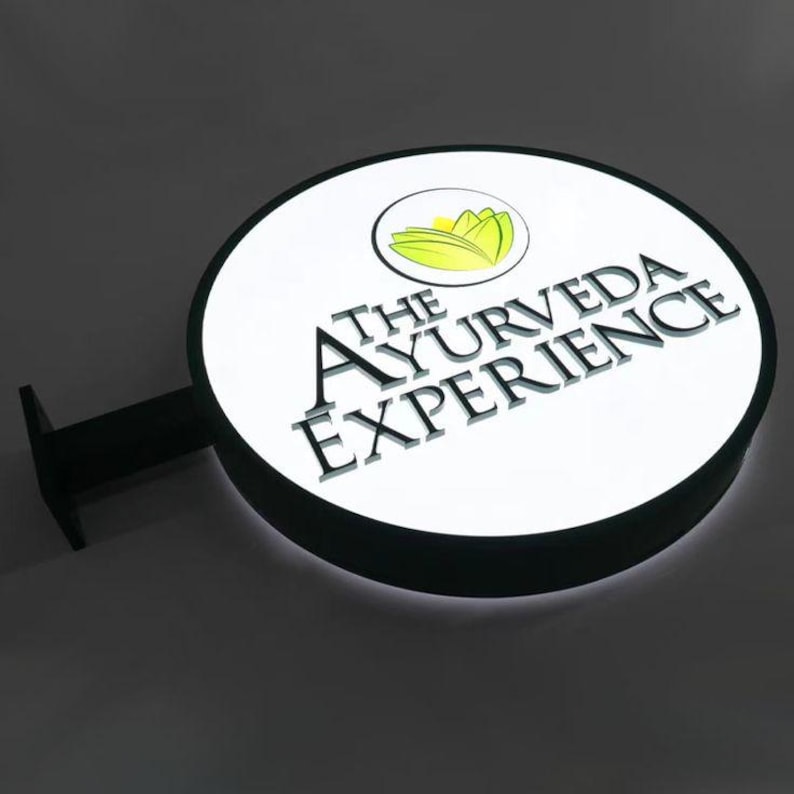K&ouml;nnte beinhalten: Ein rundes, beleuchtetes Schild mit dem Text "THE AYURVEDA EXPERIENCE" in schwarzer Schrift. Das Schild zeigt ein Logo einer gelben Blume und gr&uuml;nen Bl&auml;ttern. Das Schild ist wei&szlig; mit einem schwarzen Rand und einer schwarzen Halterung.