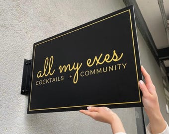 Könnte beinhalten: Schwarzes rechteckiges Schild mit goldenem Rand und Text "all my exes COCKTAILS + COMMUNITY". Das Schild ist an einer grauen Wand mit einer schwarzen Metallhalterung befestigt.