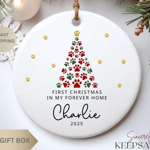 May include: White ceramic Christmas ornament with a paw print Christmas tree design in red and green. The ornament has the text "First Christmas in my forever home" and the name "Charlie" with the year 2025.