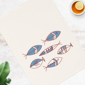 Puede incluir: Un mantel individual de lino blanco con un diseño de peces estilizados en azul y rojo. Los peces están delineados en rojo y rellenos de azul.