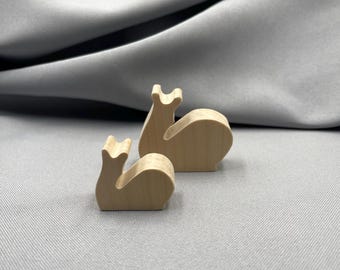 May include: Two small, wooden snail figurines. The snails are light brown and have a simple, rounded design. The background is a gray fabric.