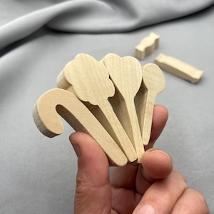 May include: A collection of unfinished wooden craft shapes, including a candy cane, flower, heart, and spoon designs. Additional small wooden pieces are visible in the background. The items are held in a hand against a gray background.