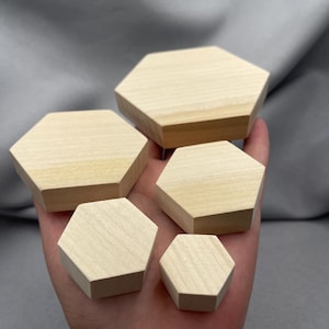 May include: A collection of five unfinished wooden hexagon shapes in varying sizes. The wood has a light, natural color with visible grain. The shapes are held in a hand, showcasing their geometric design and potential for crafts or decor.