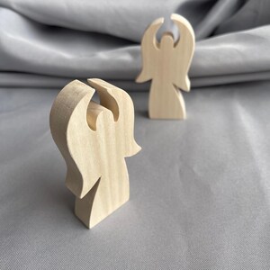 Puede incluir: Dos figuras de ángeles de madera. Los ángeles son de color claro, con un diseño sencillo. Un ángel está en primer plano y el otro en segundo plano. Los ángeles tienen un diseño recortado para la cabeza y las alas.