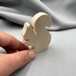 May include: A wooden squirrel figurine, carved from light-colored wood. The squirrel is depicted in a side profile, with a curved tail and detailed features. The figurine is held in a hand against a gray background.