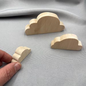 May include: Three light-colored, smooth wooden cloud-shaped cutouts of varying sizes. Each piece features a rounded, multi-lobed top edge and a flat base, allowing them to stand upright. The natural wood grain is visible on the surface of each.