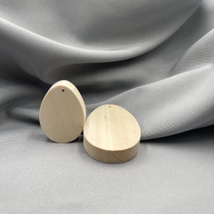 Może przedstawiać: Dwa niedokończone drewniane wisiorki w kształcie łezki. Jeden stoi pionowo, a drugi leży na boku. Każdy ma mały otwór u góry do nawlekania. Wisiorki są jasne i gładkie, z widocznym usłojeniem drewna.