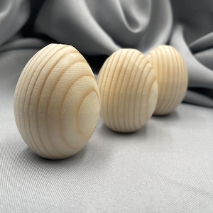 Może przedstawiać: Trzy drewniane przedmioty w kształcie jajka z widocznym usłojeniem drewna. Jajka są jasne i stoją na szarej powierzchni. Tło to miękka, szara tkanina.
