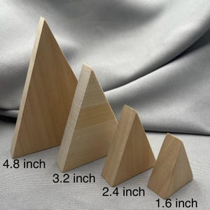 Może przedstawiać: Cztery drewniane graniastosłupy trójkątne o różnych rozmiarach. Największy ma 12,2 cm wysokości, następny 8,1 cm, potem 6,1 cm, a najmniejszy 4,1 cm. Pryzmaty wykonane są z jasnego drewna i prezentowane na szarym tle.