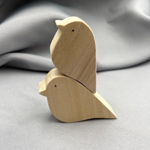 Puede incluir: Dos figuras de pájaros de madera apiladas una encima de la otra. Los pájaros son de color marrón claro con formas simples y redondeadas y pequeños detalles oscuros para los ojos. El fondo es una tela gris suave.