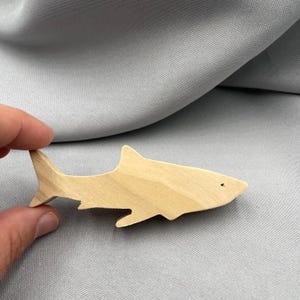 Könnte beinhalten: Holzfigur eines Hais mit natürlicher Holzoberfläche. Der Hai hat eine Rückenflosse, eine Schwanzflosse und ein kleines Augendetail. Die Figur wird vor einem grauen Stoffhintergrund gehalten.