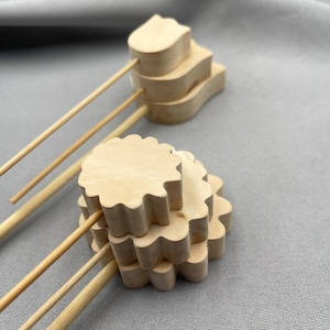 May include: Two wooden drink muddlers with decorative tops. One muddler has a layered, tulip-like design, and the other has a scalloped, flower-like design. The muddlers are made of light-colored wood and have long, thin wooden handles.