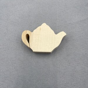 May include: A small, light-coloured wooden teapot cutout. The teapot has a handle, spout, and a lid. The wood grain is visible. The cutout is placed on a grey fabric surface.