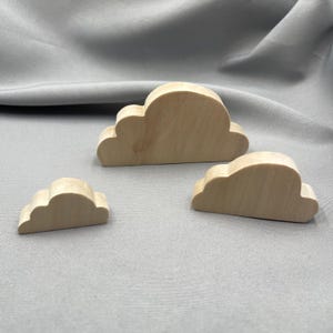 May include: Three light-coloured wooden cloud-shaped cutouts of varying sizes. Each piece features a smooth, natural wood grain finish with rounded edges, designed as flat, freestanding shapes that mimic the appearance of stylised clouds.