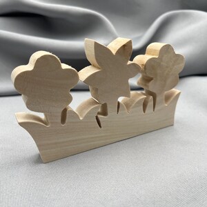 May include: A light-colored, unfinished wooden cutout featuring three distinct flower shapes emerging from a base that resembles a grassy patch. The wood has a smooth, natural grain texture and a flat, standing design with clean, laser-cut edges.