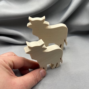 Könnte beinhalten: Zwei Holzfiguren von Kühen, eine größer als die andere, vor einem grauen Hintergrund. Die Kühe sind hell, möglicherweise unfertiges Holz, mit einfachen, blockartigen Formen. Die kleinere Kuh wird von einer Hand gehalten.