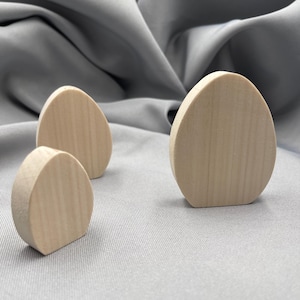 Może przedstawiać: Trzy niedokończone drewniane kształty jaj o różnych rozmiarach. Jajka są jasne i mają gładkie, naturalne usłojenie drewna. Ułożone są na szarym tle z tkaniny, sugerując motyw rzemieślniczy lub dekoracyjny.