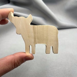 Puede incluir: Un recorte de madera con forma de vaca, sostenido por una mano. La vaca es de color marrón claro con vetas de madera visibles, con cuernos, cabeza, cuerpo y patas. El recorte es probablemente para manualidades o decoración.