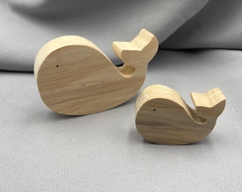 May include: Two wooden whale figurines. The larger whale is approximately 4 inches long, and the smaller whale is about 2 inches long. Both are carved from light-colored wood, with simple details like a tail and a small eye.