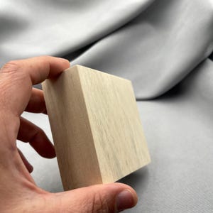 Könnte beinhalten: Ein hellfarbiger, quadratischer Holzblock mit sichtbarer Holzmaserung. Der Block wird in einer Hand gehalten und zeigt seine glatte, unfertige Oberfläche. Das Holz hat eine hellbraune Farbe.