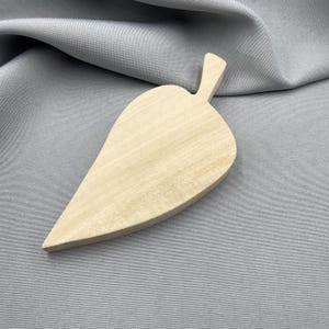 May include: A light-coloured, smooth wooden object shaped like a leaf with a pointed tip and a small stem at the top. The wood grain is visible across the surface, showing natural variations in the light beige tone of the material.