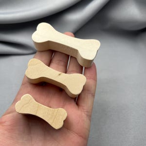 May include: Three wooden dog bone shapes in varying sizes. The bones are light-colored, with a natural wood grain. The shapes are held in a hand against a gray background.