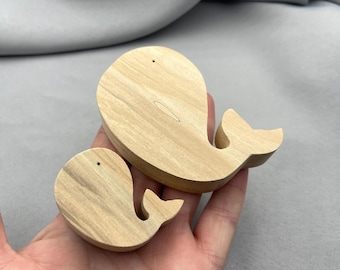 May include: Two wooden whale-shaped ornaments. The whales are light-colored, with visible wood grain. One whale is larger than the other. Each whale has a small hole near the top and a curved tail. The whales are resting on a gray surface.