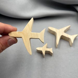 以下が含まれることがあります： さまざまなサイズの木製飛行機の切り抜きが3つ。 飛行機は明るい色の木材で作られており、シンプルなデザインです。 飛行機は手で持たれています。 背景は柔らかな灰色の生地です。