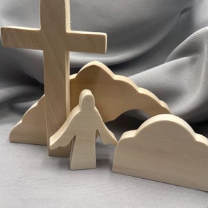 以下が含まれることがあります： 十字架、腕を広げた人物、雲の形をした要素など、木製の宗教的なフィギュアのセット。これらのフィギュアは明るい色の木材で作られ、灰色の背景に配置されています。