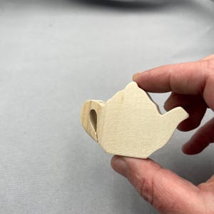 May include: A small, unfinished wooden teapot. The teapot is held between fingers and features a handle, spout, and lid, all carved from light-coloured wood. The design is simple and rustic, suggesting a craft or DIY project.