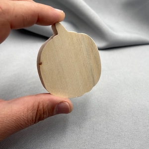 Könnte beinhalten: Ein heller, unbehandelter Holzausschnitt in Form eines Kürbisses. Die Oberfläche zeigt natürliche Holzmaserungen und eine glatte, flache Textur. Die Kürbisform weist einen kleinen, spitzen Stiel an der Oberseite und einen abgerundeten Körper auf.