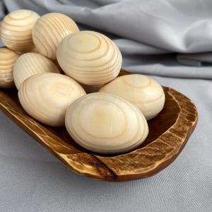 Könnte beinhalten: Eine Sammlung unfertiger Holzeier mit sichtbarer Maserung, präsentiert in einer rustikalen, braunen Holzschale. Die Eier variieren in Größe und Form und haben eine natürliche, helle Holzfarbe. Die Schale hat ein geschnitztes, strukturiertes Aussehen, vor grauem Hintergrund.