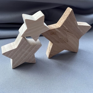 Può includere: Tre decorazioni a forma di stella in legno di varie dimensioni. Le stelle sono realizzate in legno di colore chiaro, con motivi di venature visibili. Le stelle sono disposte in un gruppo, con una stella che si sovrappone leggermente alle altre. Lo sfondo è di colore grigio.