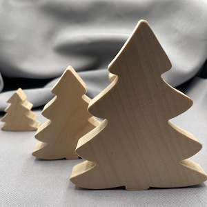 May include: Three light-colored wooden tree cutouts of varying sizes arranged in a row. Each tree features a simple, stylized silhouette with tiered branches and a flat base, showcasing the natural grain and smooth texture of the light wood.