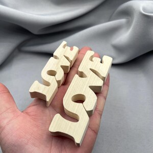 May include: Two wooden "LOVE" word cutouts in a light, natural wood tone. The letters are separate and held in a hand, with a soft grey background.