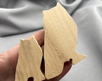 May include: Two wooden cat figurines. The larger cat is approximately 4 inches tall, and the smaller cat is about 2 inches tall. Both are cut from light-colored wood, with simple, stylized shapes. The cats are positioned close together, suggesting a parent and child.