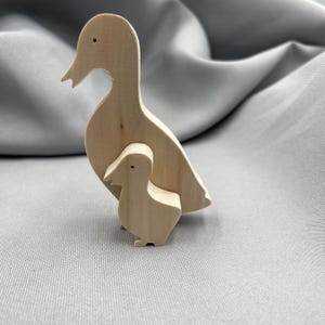 May include: A wooden duck and duckling figurine set. The larger duck is standing with its wings spread out, and the smaller duckling is nestled under its mother's wing. Both ducks are made of natural wood and have a simple, rustic design.